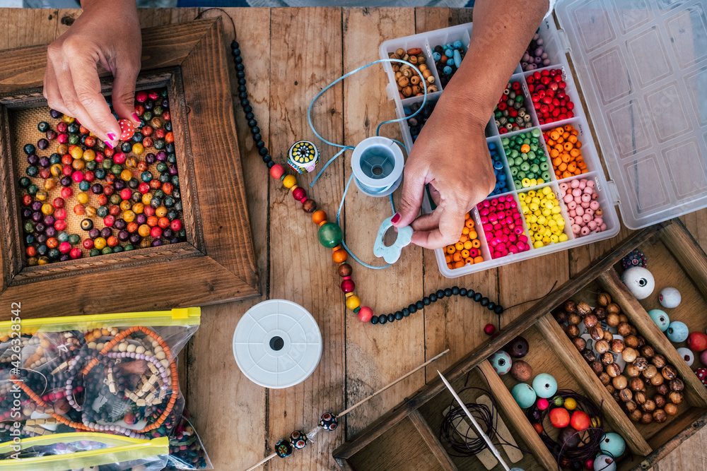 A person is making a beaded bracelet at a wooden table. There are various beads, cord, and tools scattered around.